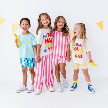 Four children standing together in a bright studio setting, each wearing a colorful graphic T-shirt and matching shorts or pants. The outfits feature playful prints and patterns including blue/yellow, pink/white stripes, and cream/yellow graphics.
