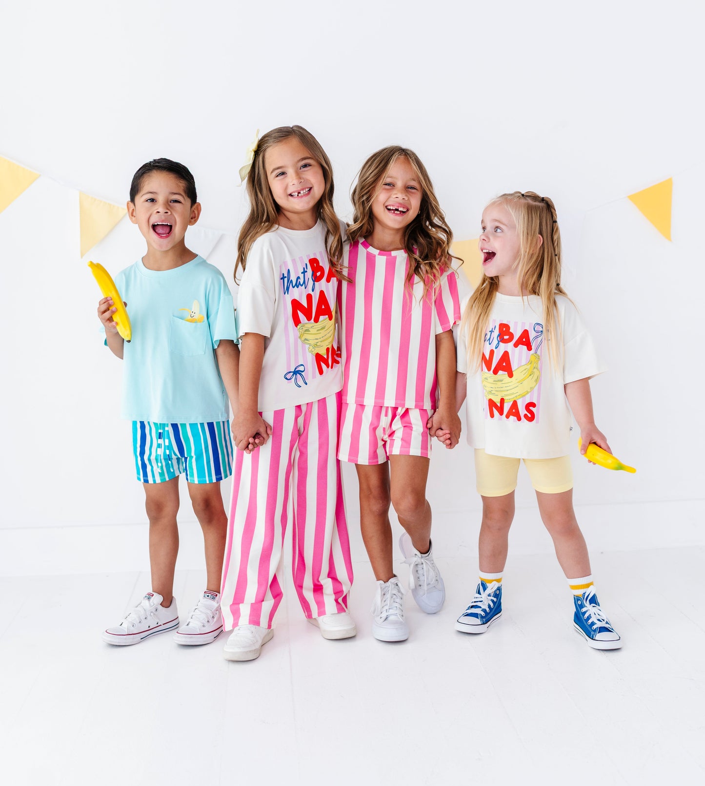 Four children standing together in a bright studio setting, each wearing a colorful graphic T-shirt and matching shorts or pants. The outfits feature playful prints and patterns including blue/yellow, pink/white stripes, and cream/yellow graphics.
