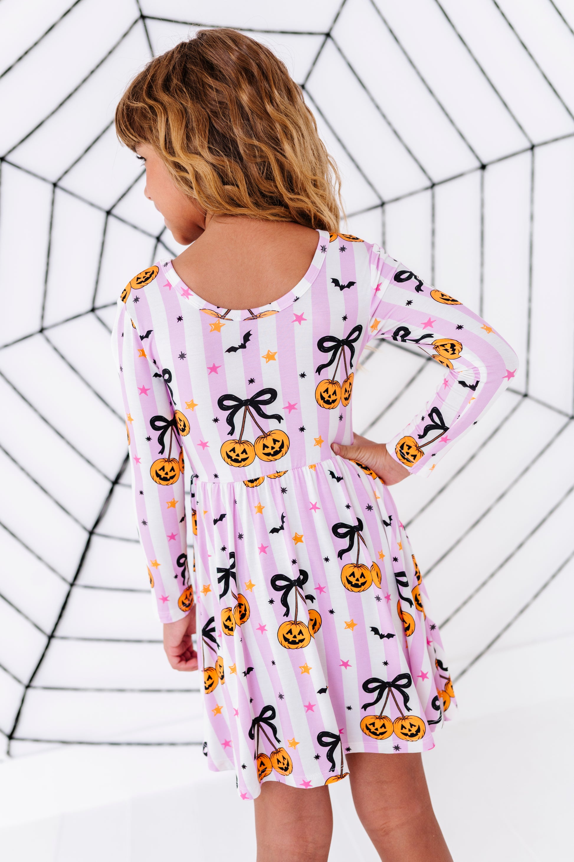 A young girl with blonde hair is shown from behind wearing a pink and white striped long-sleeve dress with a Halloween print of pumpkins, black bows, and bats. She is standing against a white background with a black spiderweb pattern.