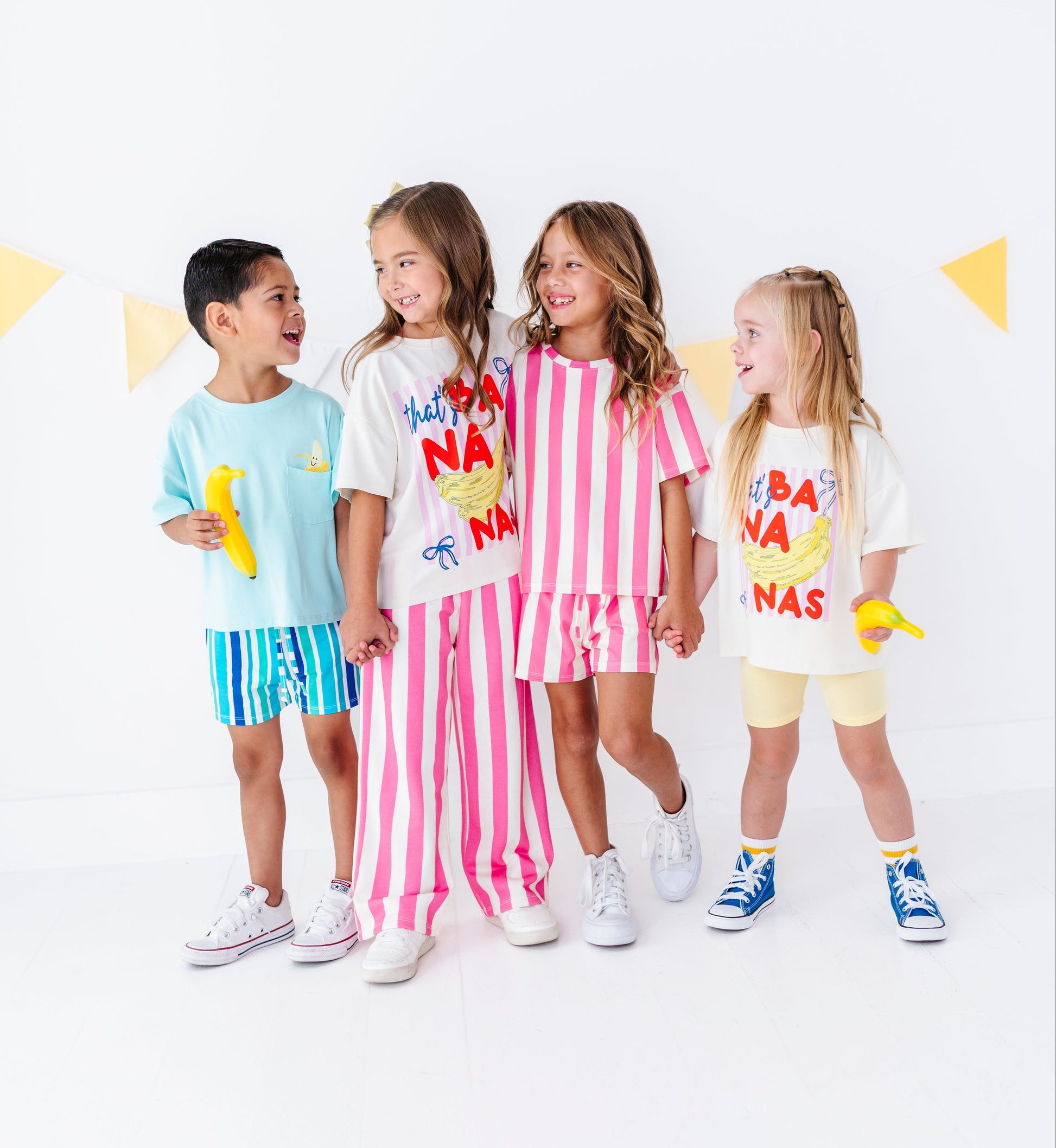 Four children modeling matching graphic t-shirts and shorts sets in a bright studio setting. The outfits feature playful banana-themed graphics and color-blocked patterns, including light blue, white with pink stripes, and cream with yellow accents.
