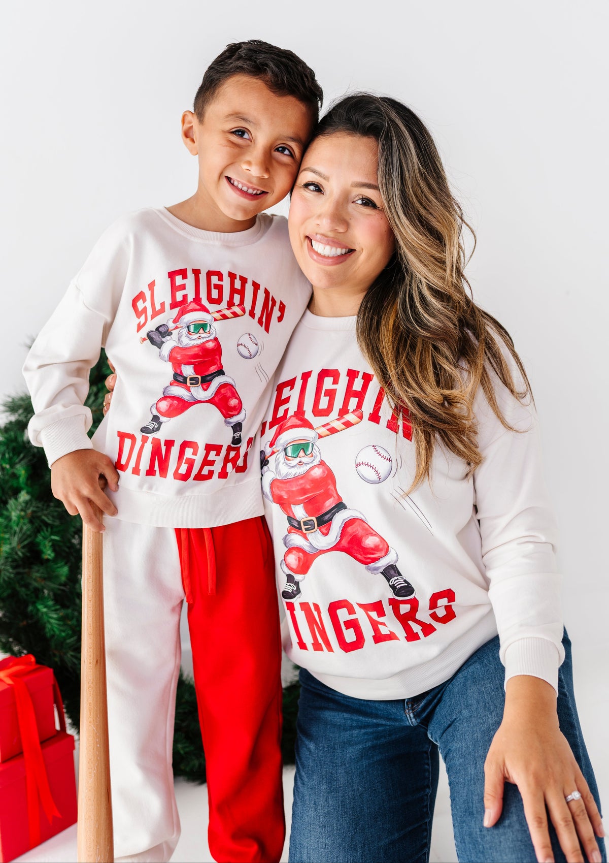 A smiling man and woman pose together wearing matching white long-sleeve sweatshirts with a red and black sleigh graphic and the words 'Sleigh Dinger.' The man also wears red sweatpants, and the woman wears blue jeans.