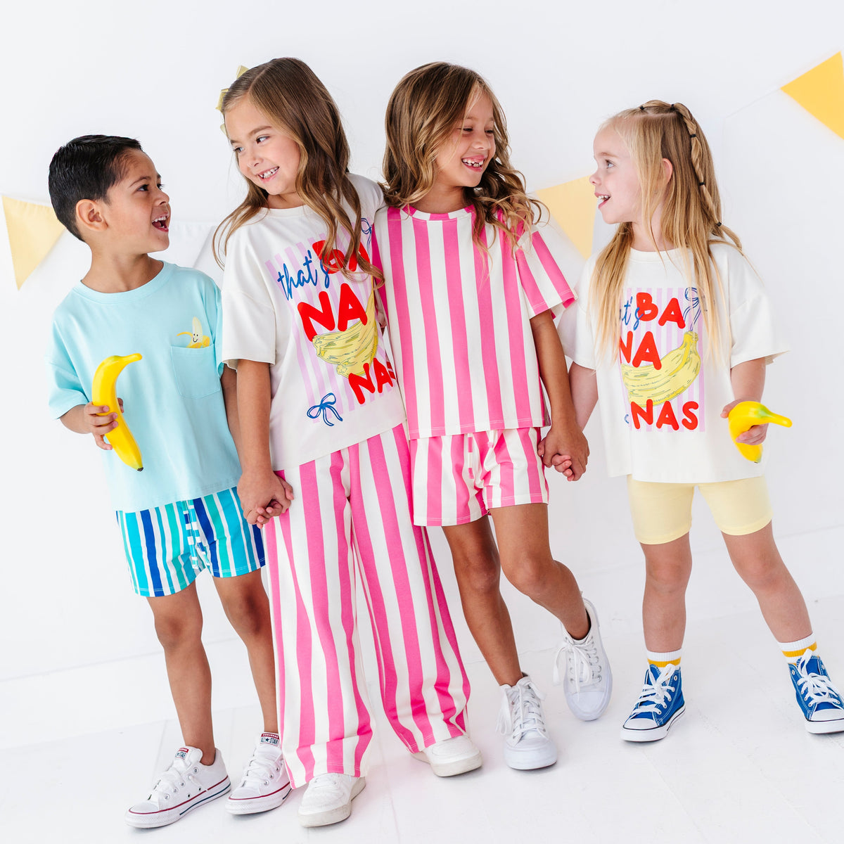 Four children modeling matching banana-themed two-piece outfits in a bright studio setting. The outfits include a light blue set, a white set with pink vertical stripes, and a white set with yellow shorts, each featuring a banana graphic and the text 'HAPPY BANANAS'.