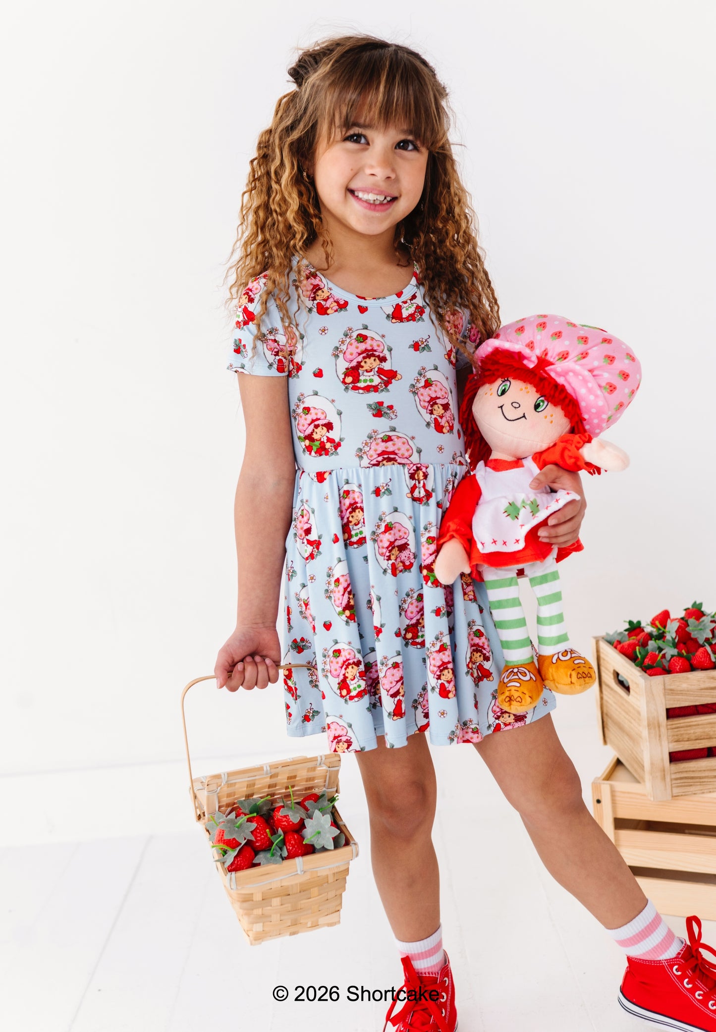 Child wearing a Strawberry Shortcake™ dress holding a Strawberry Shortcake™ doll and basket with strawberries on a white background