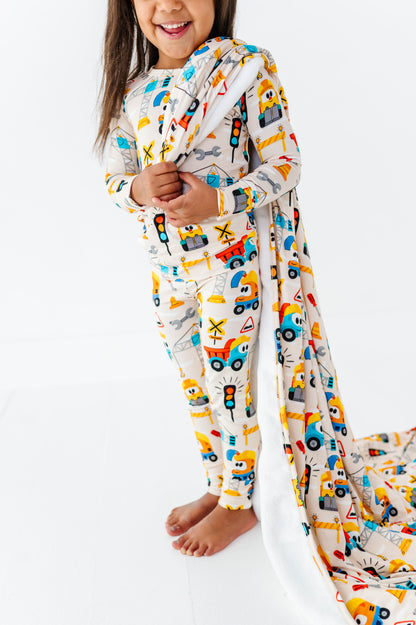 A young child wearing a long-sleeve one-piece pajama set with a colorful construction vehicle print, standing barefoot on a white studio floor while pulling a matching long blanket behind them.