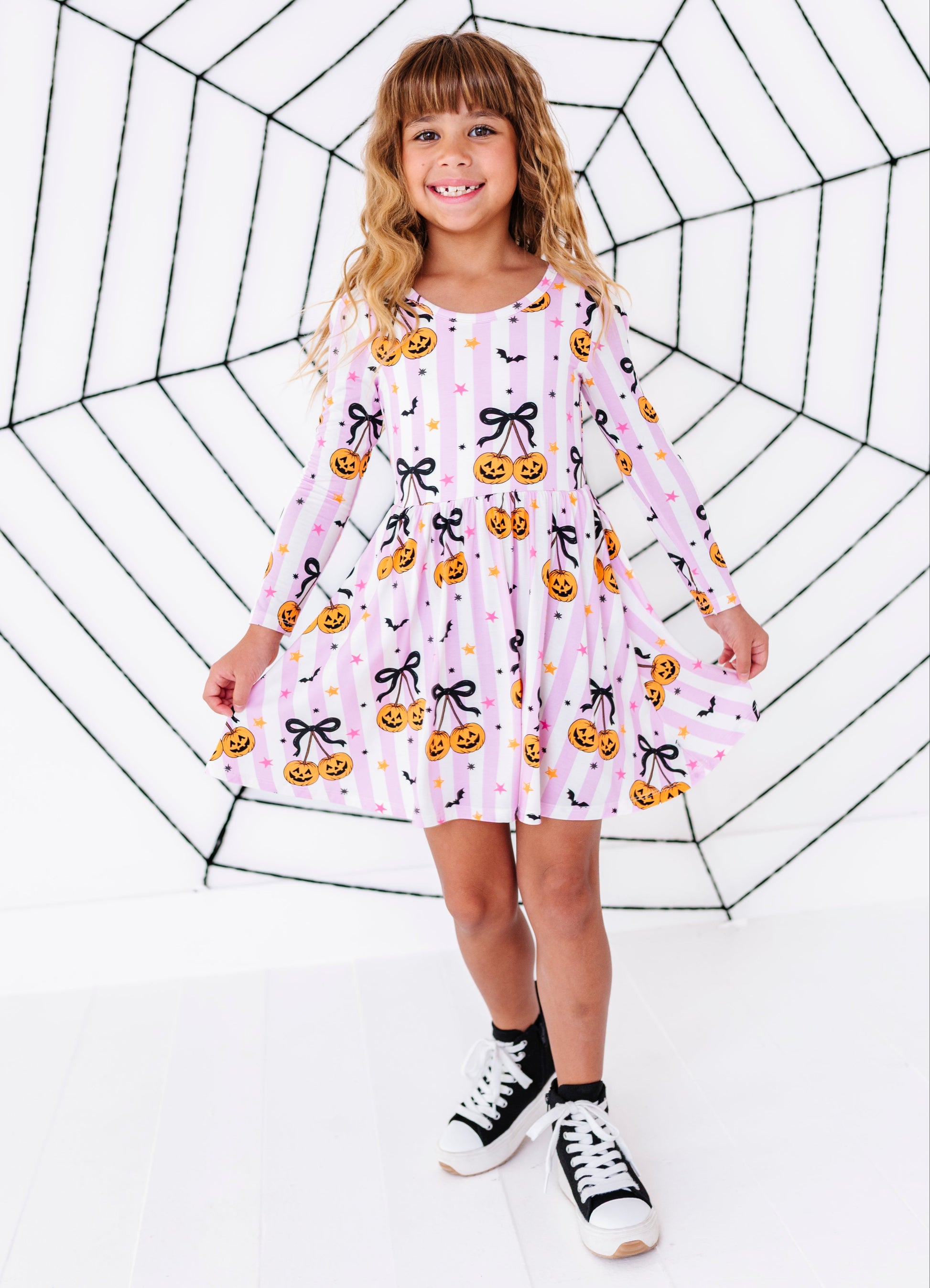 A young girl with blonde hair stands in front of a large black spiderweb graphic on a white background, wearing a pink long-sleeve dress with a pumpkin and bow pattern, black socks, and black-and-white sneakers.