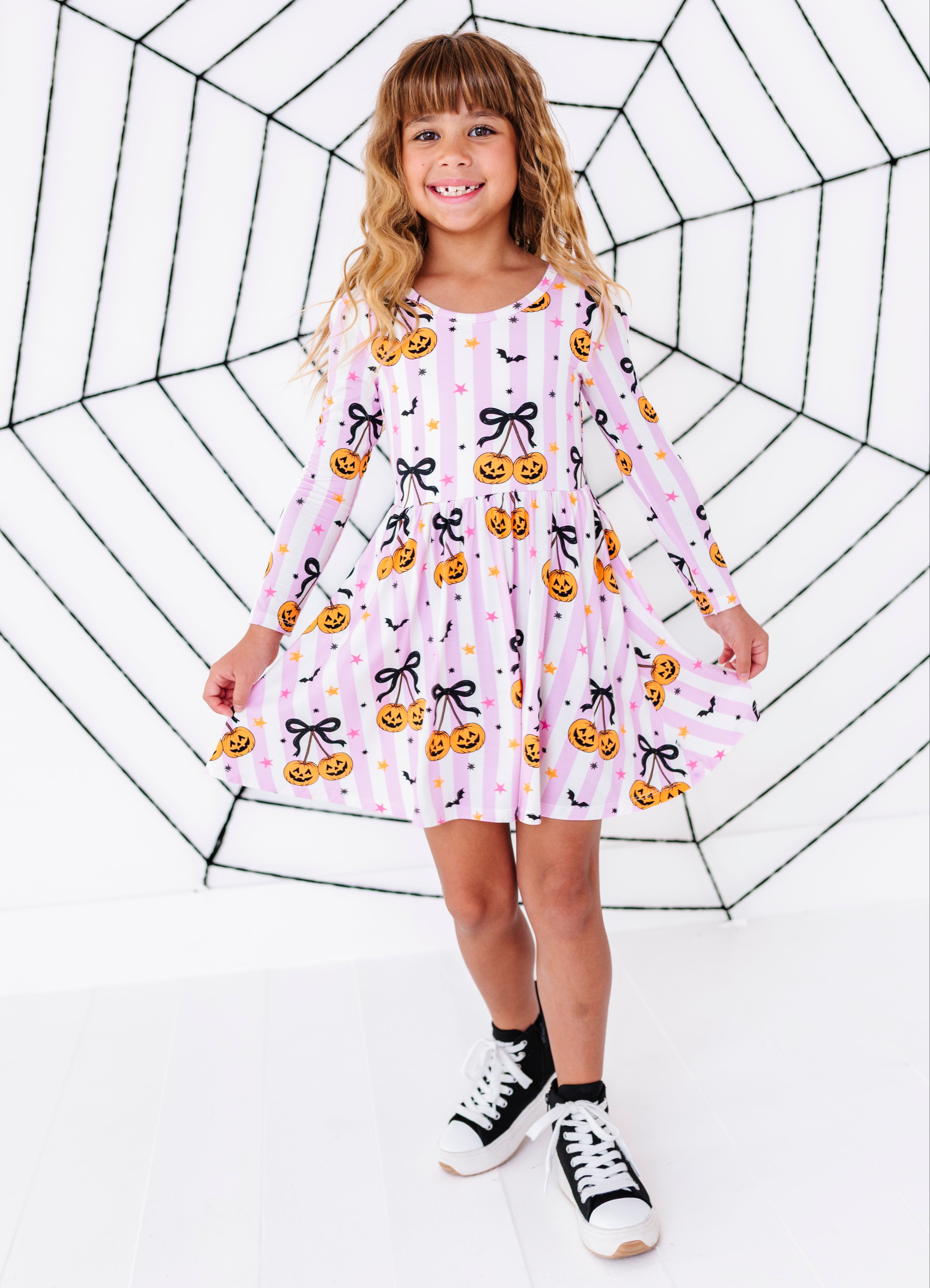 A young girl with blonde hair stands in front of a large black spiderweb graphic on a white background, wearing a pink long-sleeve dress with a pumpkin and bow pattern, black socks, and black-and-white sneakers.