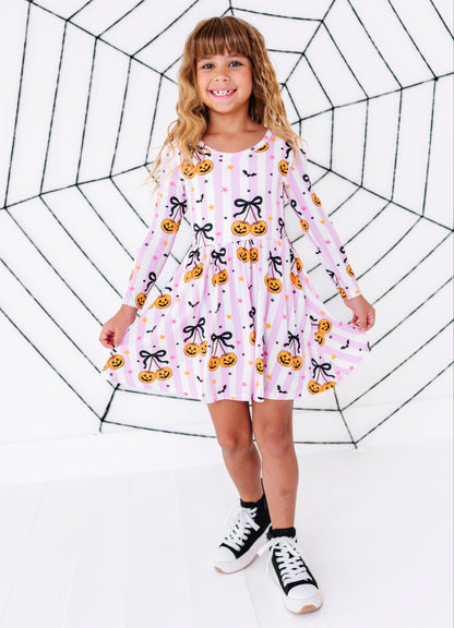 A young girl with blonde hair stands in front of a large black spiderweb graphic on a white background, wearing a pink long-sleeve dress with a pumpkin and bow pattern, black socks, and black-and-white sneakers.