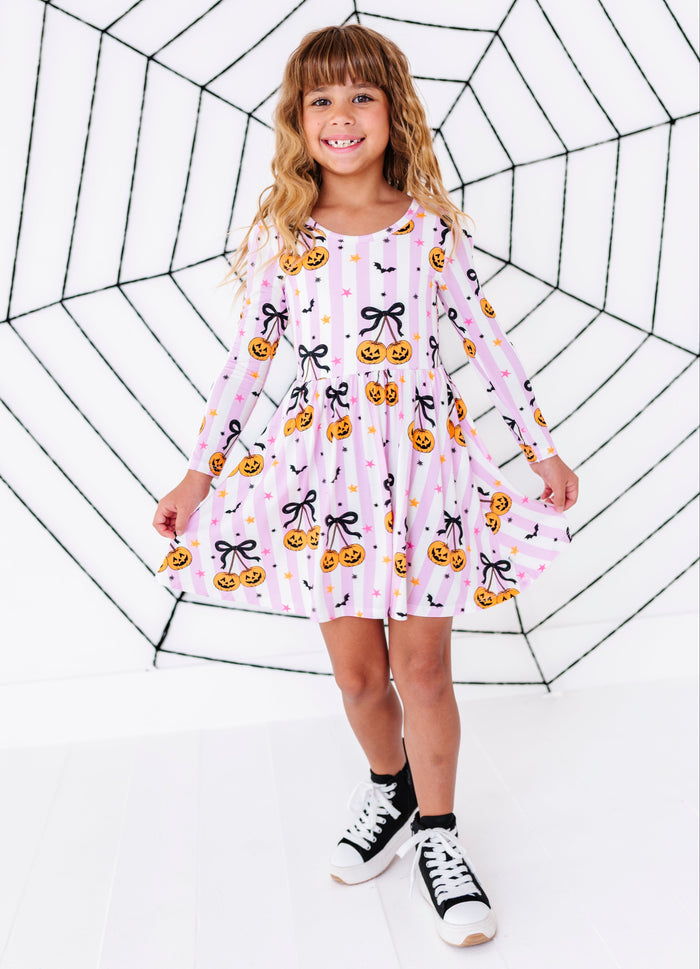 A young girl with blonde hair stands in front of a large black spiderweb graphic on a white background, wearing a pink long-sleeve dress with a pumpkin and bow pattern, black socks, and black-and-white sneakers.
