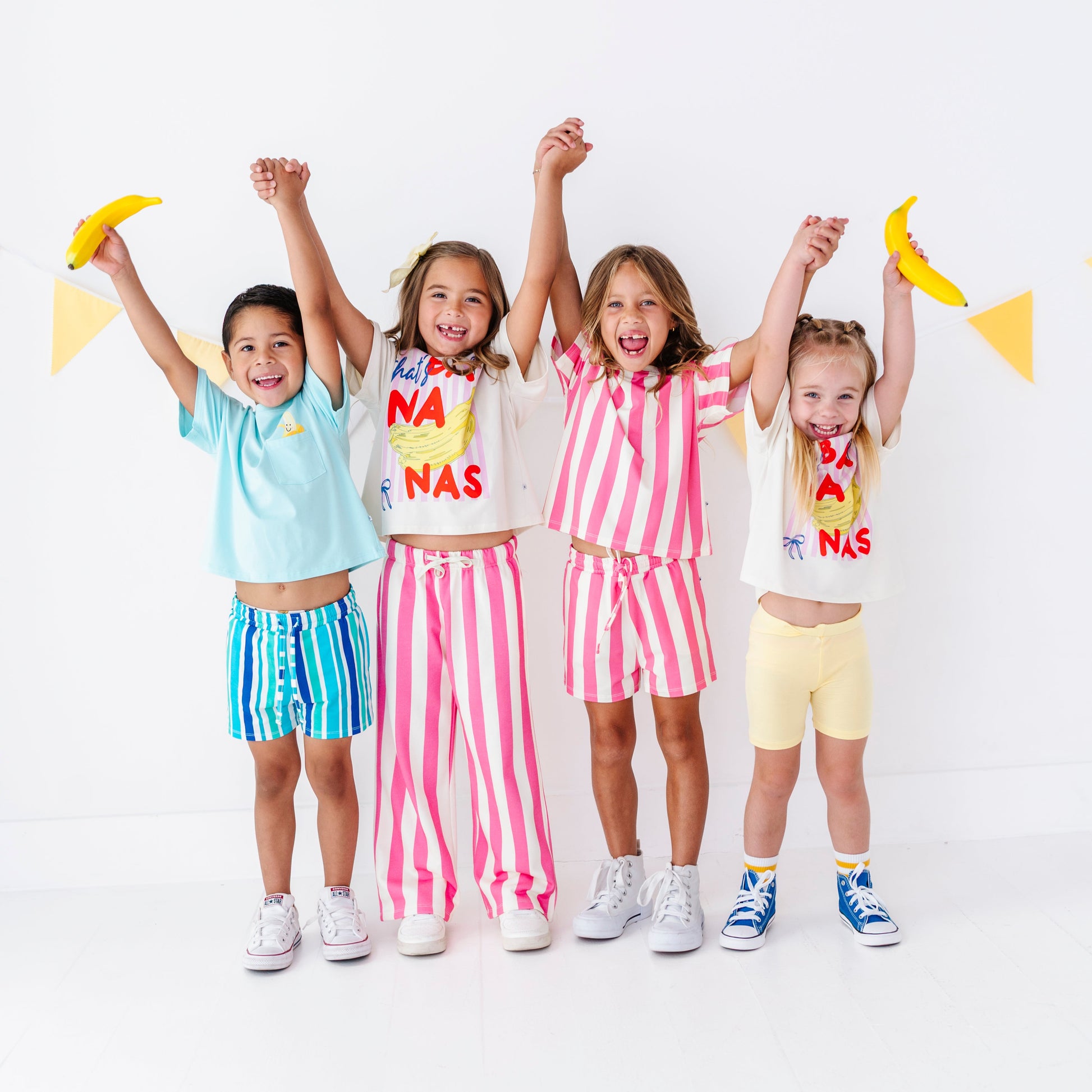 Four young girls standing together and holding hands, wearing matching coordinated outfits in pastel and bright colors. Each girl has a white T-shirt with colorful text and a matching striped or solid shorts set.