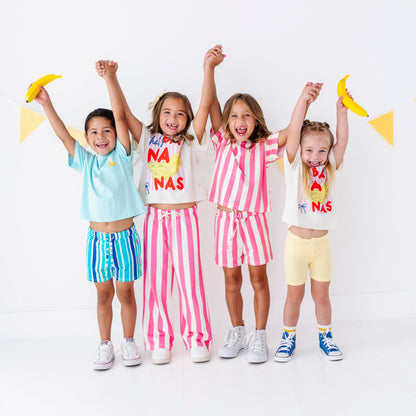 Four young girls standing together and holding hands, wearing matching coordinated outfits in pastel and bright colors. Each girl has a white T-shirt with colorful text and a matching striped or solid shorts set.