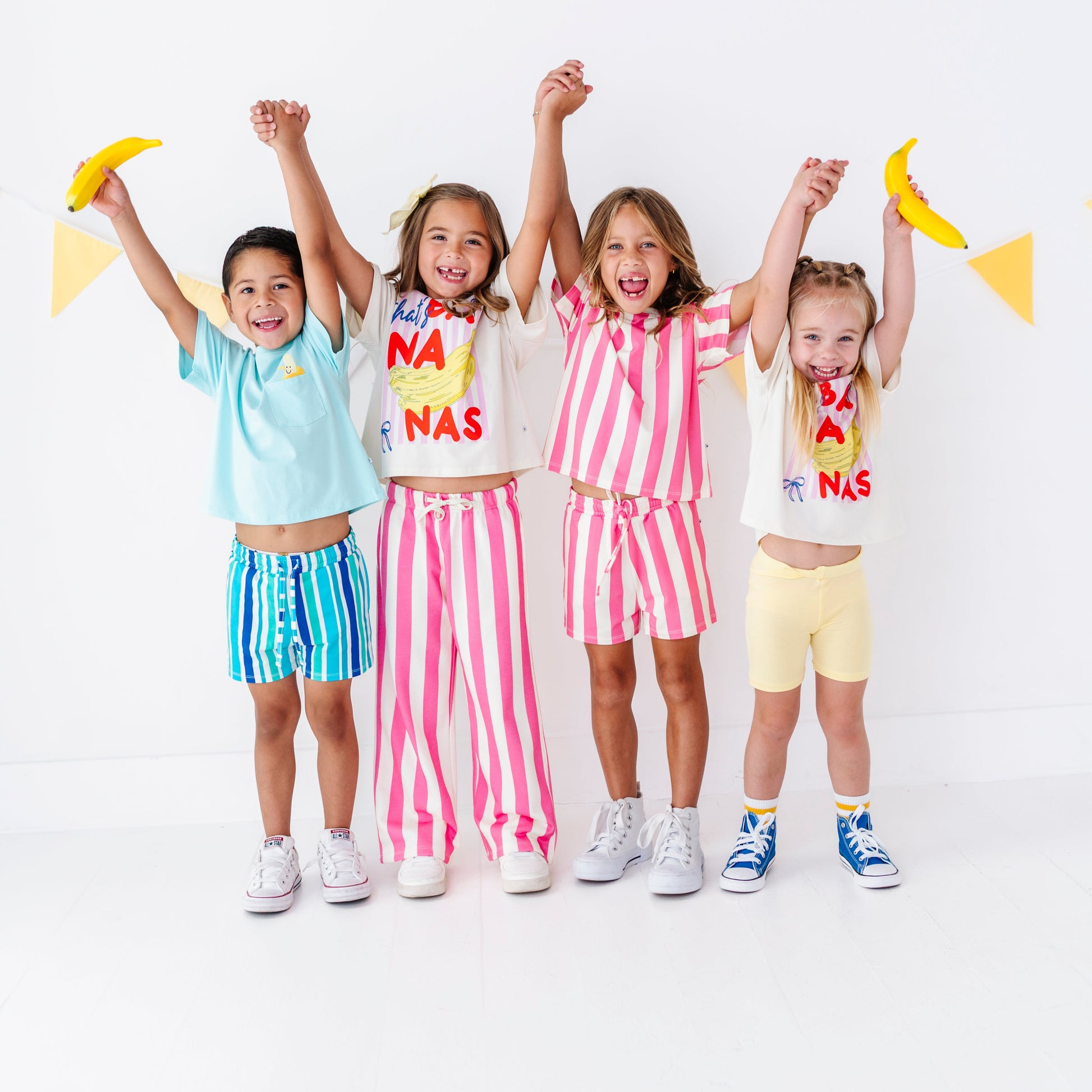 Four young kids standing together and holding hands, wearing matching coordinated outfits in pastel and bright colors. Each girl has a white T-shirt with colorful text and a matching striped or solid shorts set.