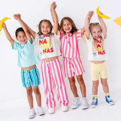 Four children standing together in a studio wearing colorful matching outfits and holding yellow banana props, with a white background.