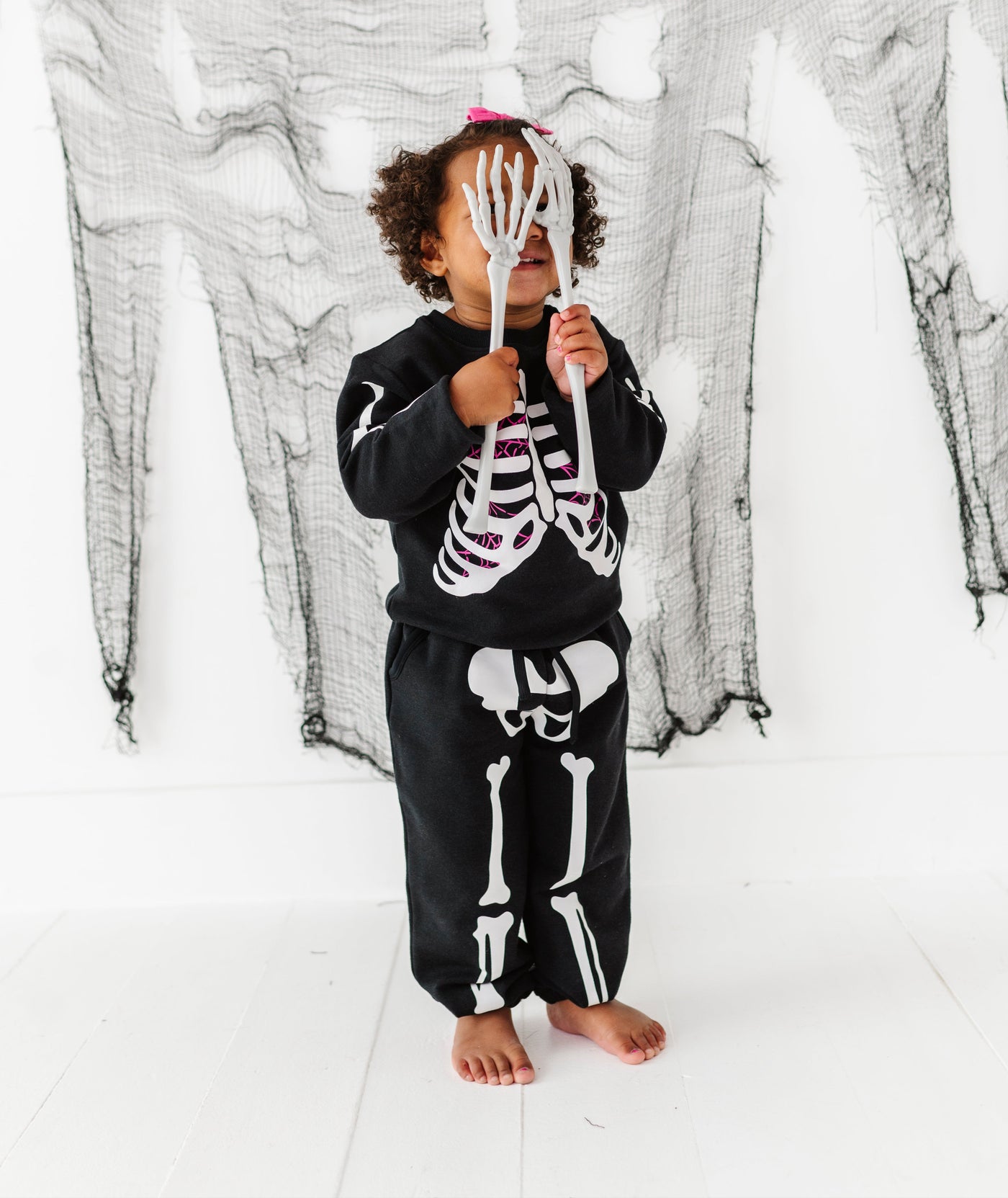 Child wearing a skeleton-themed outfit holding a small skeleton figure against a white background with black draped fabric.