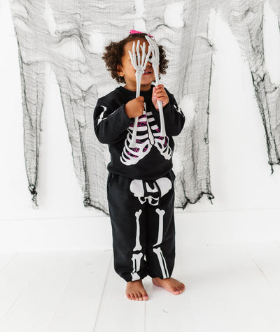 Child wearing a skeleton-themed outfit holding a small skeleton figure against a white background with black draped fabric.