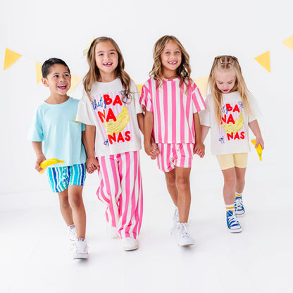 Four children walking hand-in-hand in a studio setting, wearing matching white graphic t-shirts and colorful shorts or pants sets. The shirts feature a yellow banana illustration and the text 'HAPPY BANA NAS'.