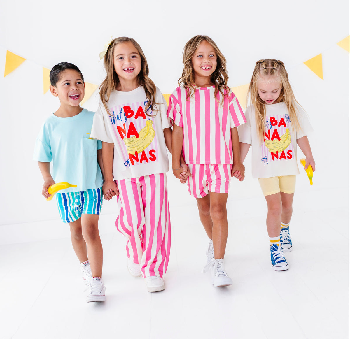 Four children walking hand-in-hand in a studio setting, wearing matching white graphic t-shirts and colorful shorts or pants sets. The shirts feature a yellow banana illustration and the text 'HAPPY BANA NAS'.