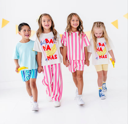 Four children walking hand-in-hand in a studio setting, wearing matching white graphic t-shirts and colorful shorts or pants sets. The shirts feature a yellow banana illustration and the text 'HAPPY BANA NAS'.