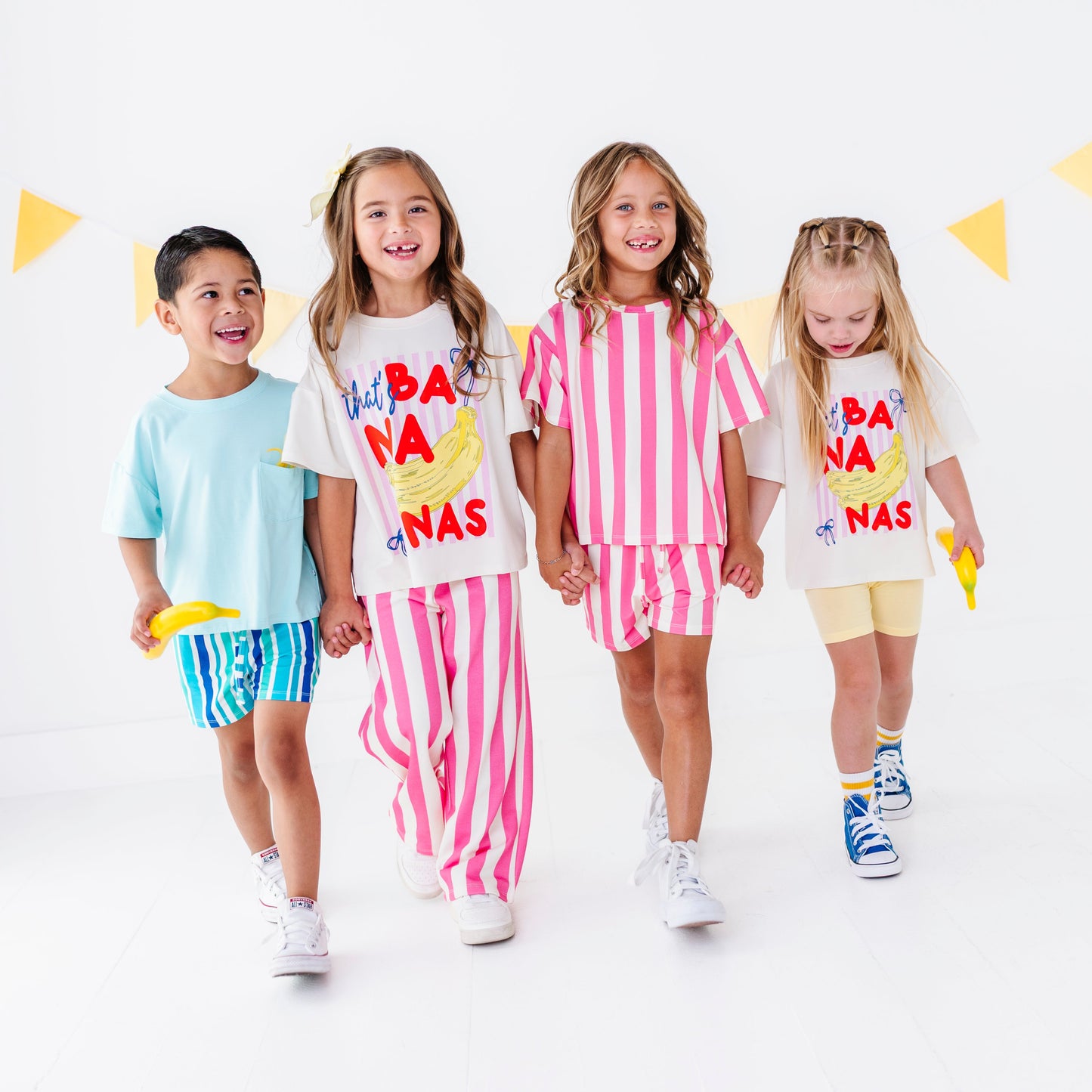 Four children walking hand-in-hand in a studio setting, wearing matching white graphic t-shirts and colorful shorts or pants sets. The shirts feature a yellow banana illustration and the text 'HAPPY BANA NAS'.