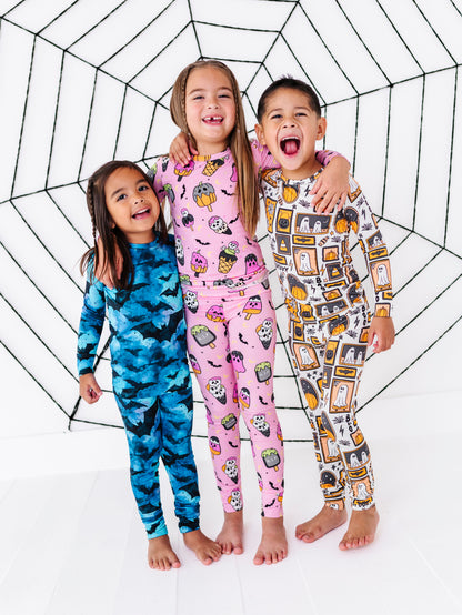 Three children wearing colorful Halloween-themed long-sleeve pajamas stand together in front of a large black spiderweb backdrop. The pajama designs feature spooky patterns including ghosts, bats, pumpkins, and haunted houses in pink, blue, and gray/orange.