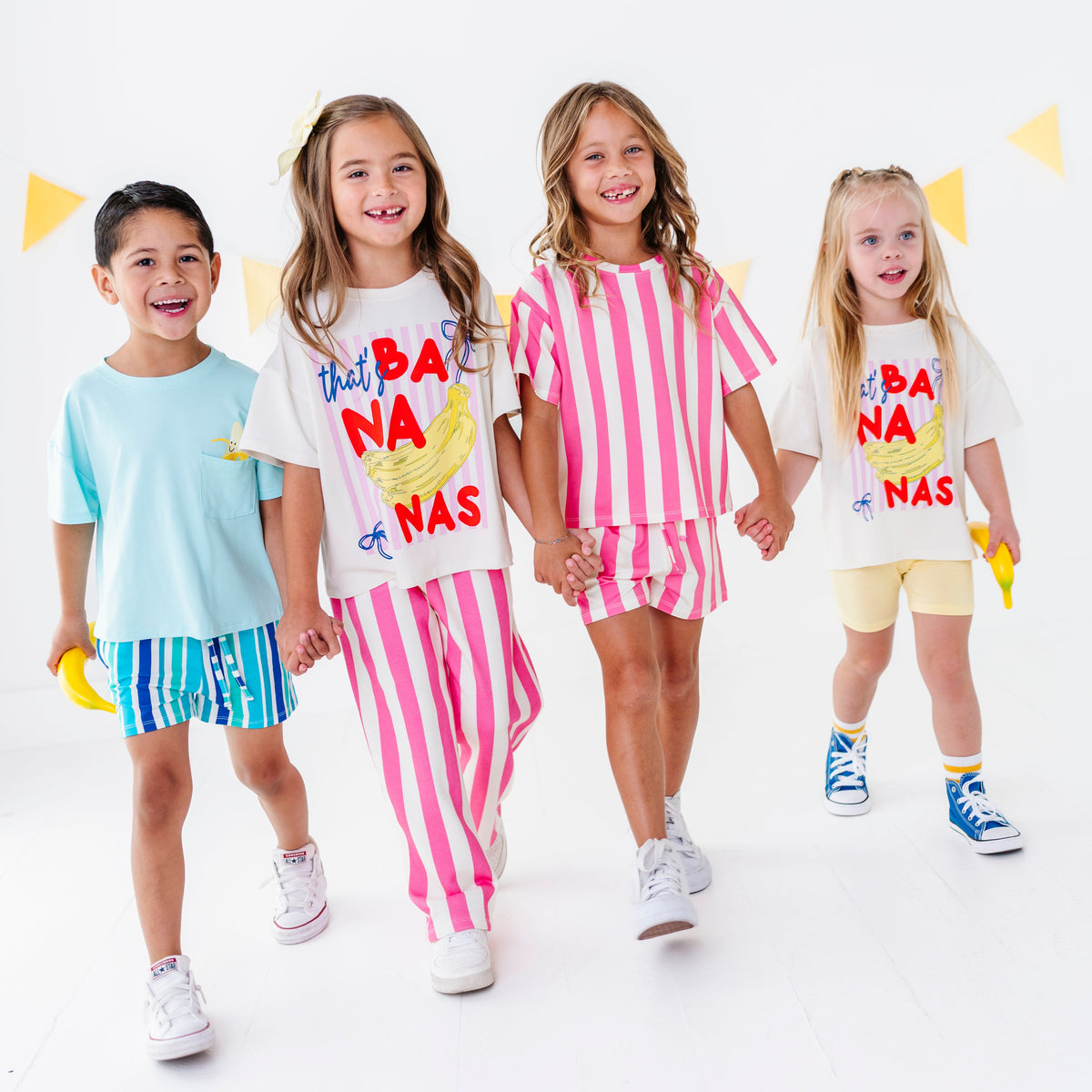 Four children modeling matching white graphic T-shirts with colorful 'Not A Banana' designs, paired with striped shorts or solid shorts, against a clean white studio background with yellow bunting.