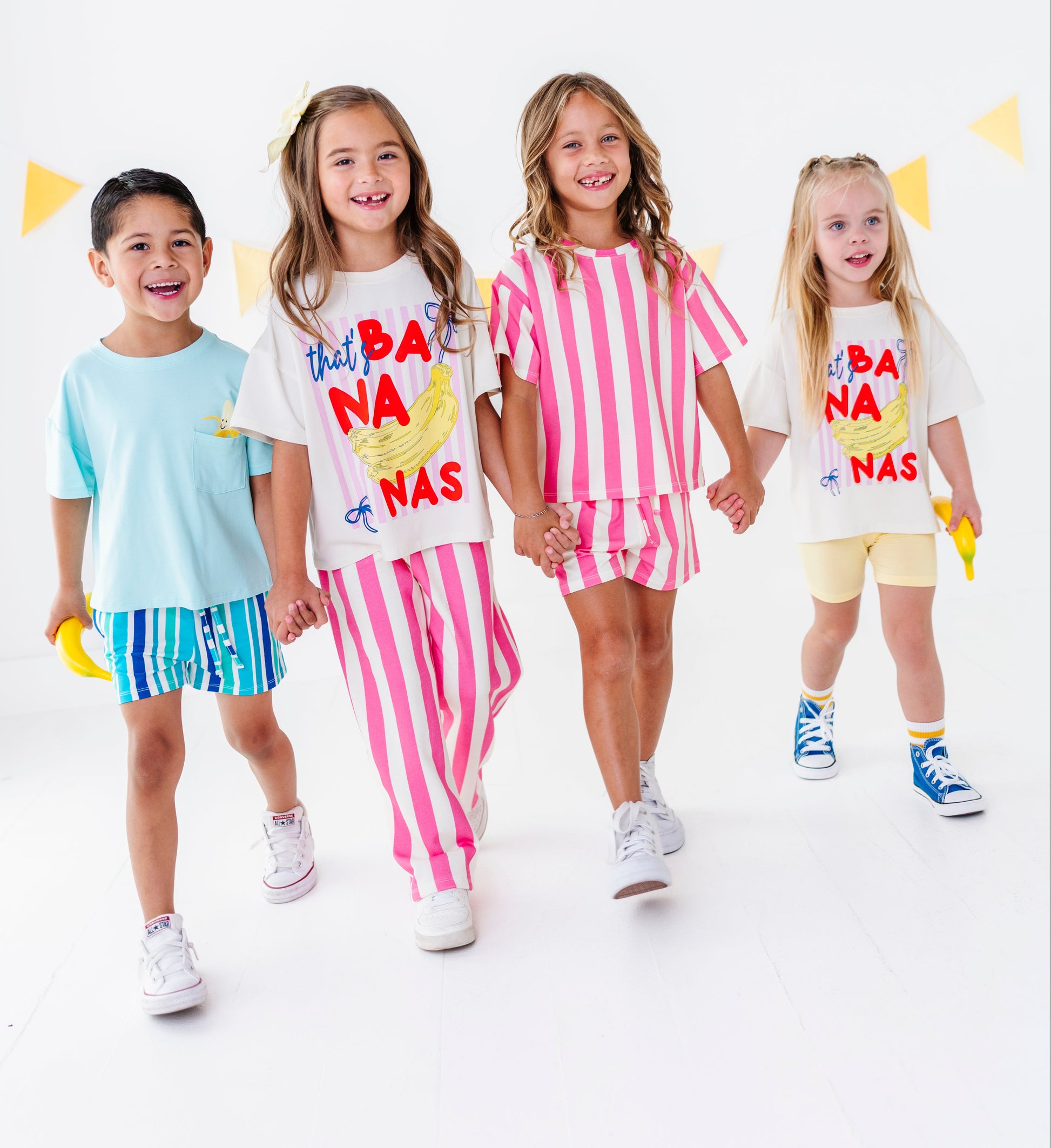 Four children modeling matching white graphic T-shirts with colorful 'Not A Banana' designs, paired with striped shorts or solid shorts, against a clean white studio background with yellow bunting.