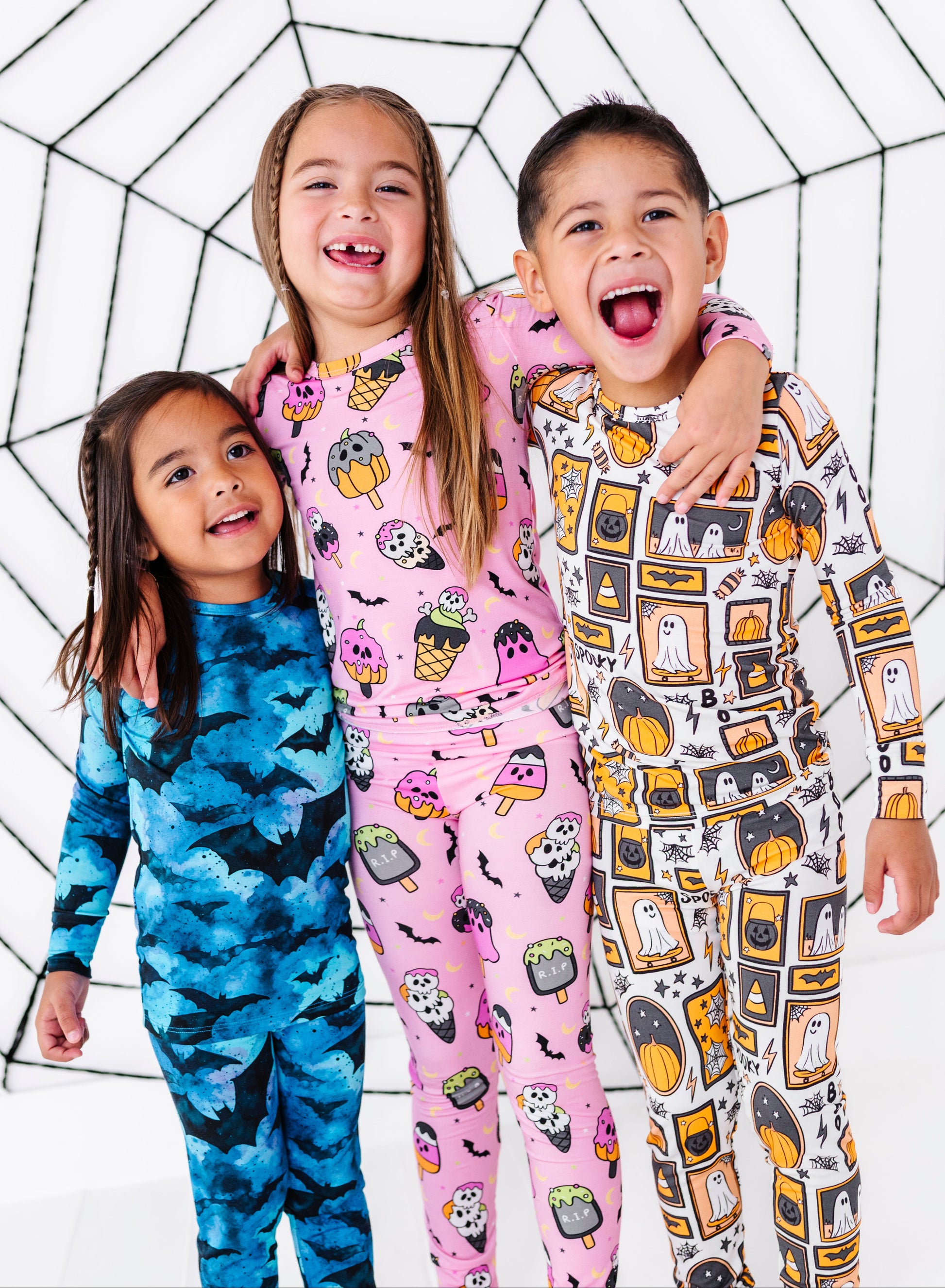 Three children wearing matching Halloween-themed long-sleeve pajamas, standing together against a white background with a black spiderweb pattern. The child on the left wears a blue pajama set with a bat pattern, the middle child wears a pink pajama set with a ghost and pumpkin pattern, and the child on the right wears a white pajama set with a ghost and pumpkin pattern.