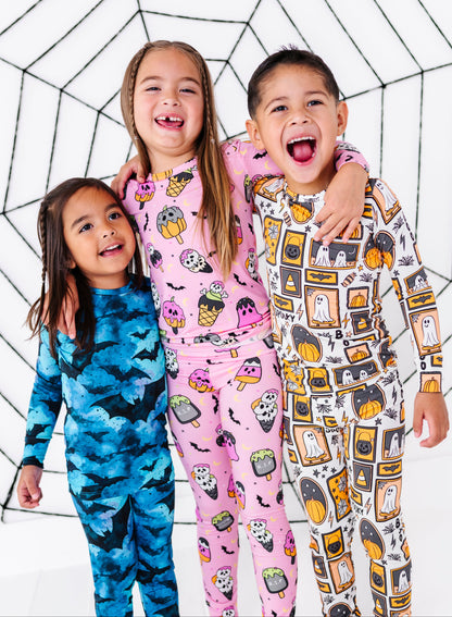 Three children wearing matching Halloween-themed long-sleeve pajamas, standing together against a white background with a black spiderweb pattern. The child on the left wears a blue pajama set with a bat pattern, the middle child wears a pink pajama set with a ghost and pumpkin pattern, and the child on the right wears a white pajama set with a ghost and pumpkin pattern.
