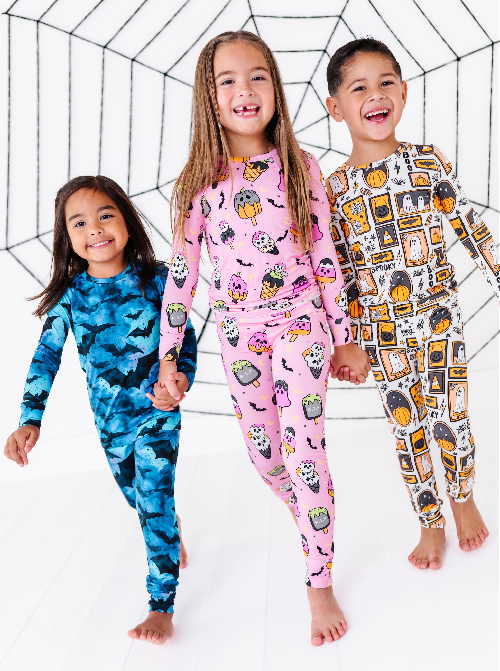 Three children wearing matching Halloween-themed long-sleeve pajamas pose together in front of a white studio backdrop with a black spiderweb graphic. The child on the left wears a blue camouflage pajama set, the center child wears a pink pajama set with a pumpkin and spider print, and the child on the right wears a white pajama set with an orange and yellow pumpkin pattern.
