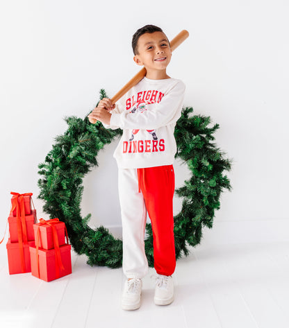 A young boy stands holding a wooden baseball bat in front of a green Christmas wreath, wearing a white long-sleeve sweatshirt with red 'SLEIGHTY DANGERS' text, white sweatpants with one leg in red, and white sneakers. Red gift boxes sit to the left on a white studio floor against a white wall.