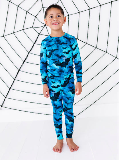 A young boy stands barefoot in front of a white studio backdrop with a large black spiderweb graphic, wearing a long-sleeve one-piece swimsuit or pajama-style top with a blue and black bat camouflage pattern.