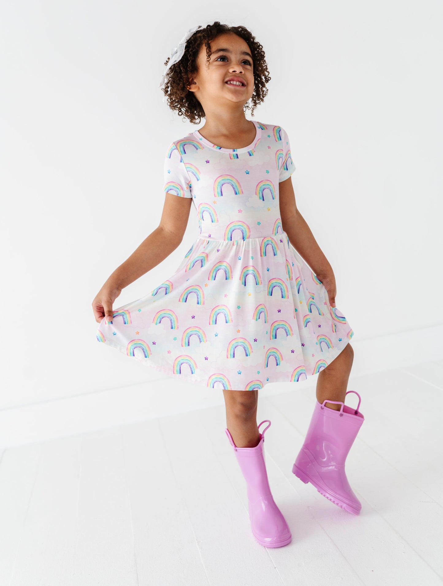 Child wearing a watercolor rainbow-patterned bamboo dress and purple rain boots on a white background