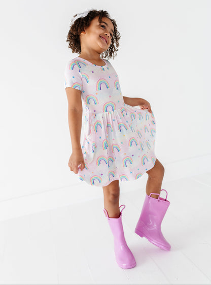 Child wearing a bamboo dress with watercolor rainbow pattern and purple rain boots on a white background
