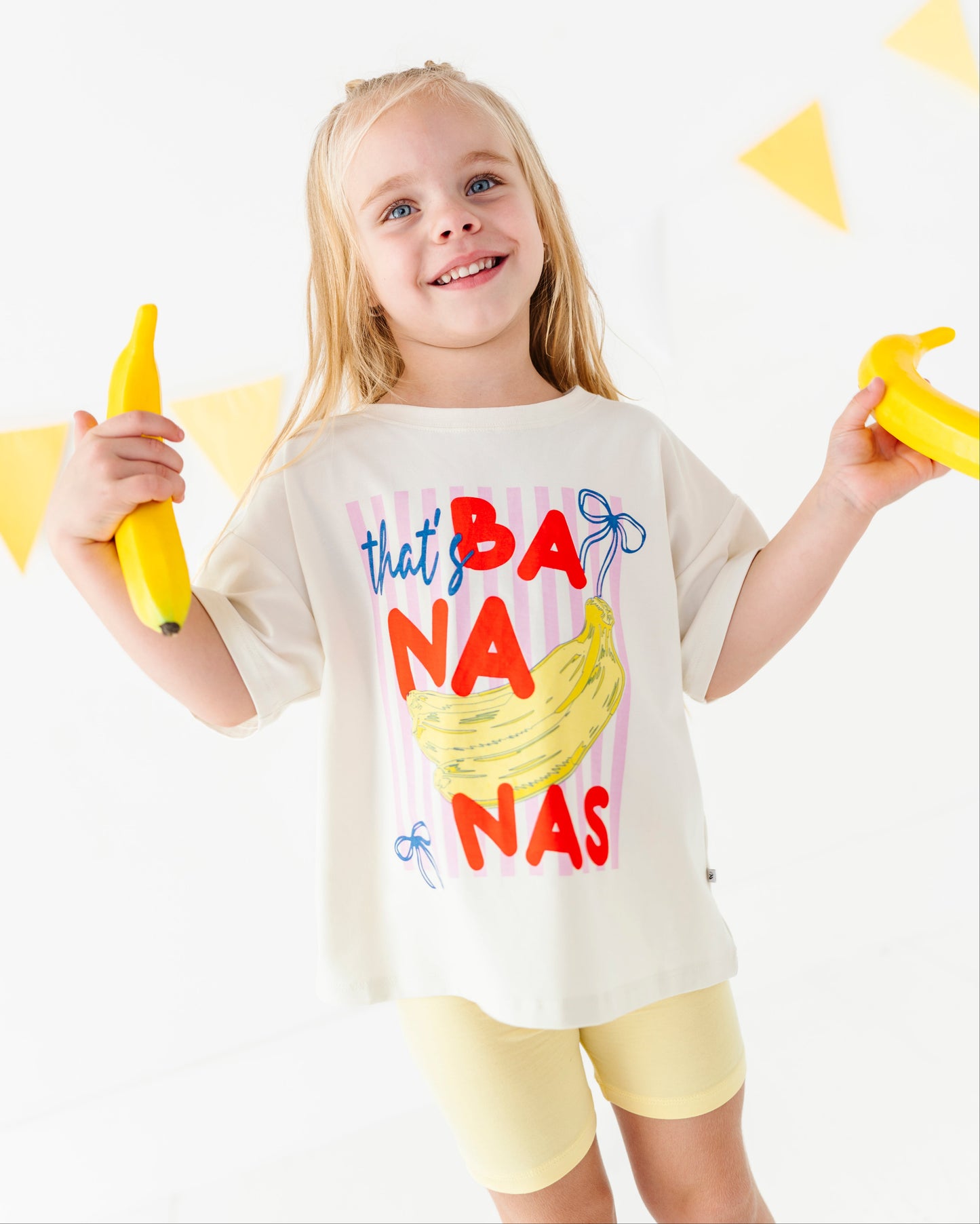 A young girl with blonde hair smiling and holding two yellow bananas, wearing a cream-colored graphic t-shirt that reads 'that's BANANAS' in red, blue, and yellow lettering, paired with light yellow shorts, against a white background with yellow triangular flags.