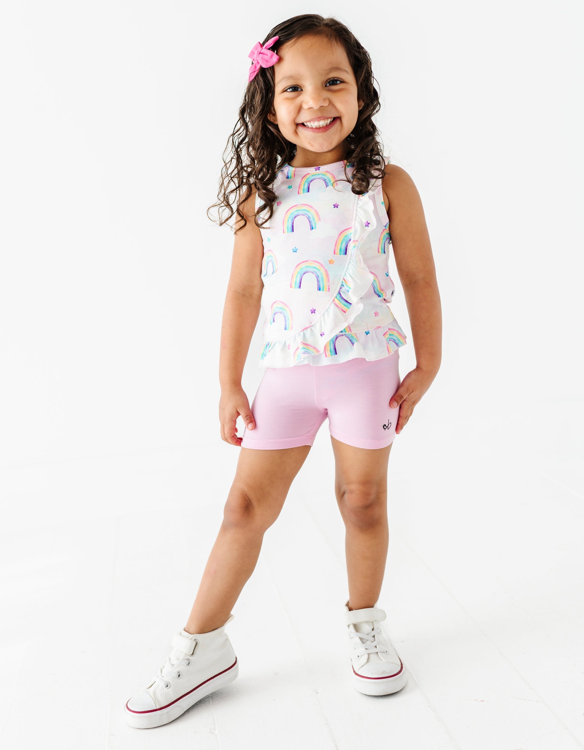 Young girl wearing a sleeveless rainbow-patterned bamboo cotton peplum top and pink bamboo biker shorts on a white background