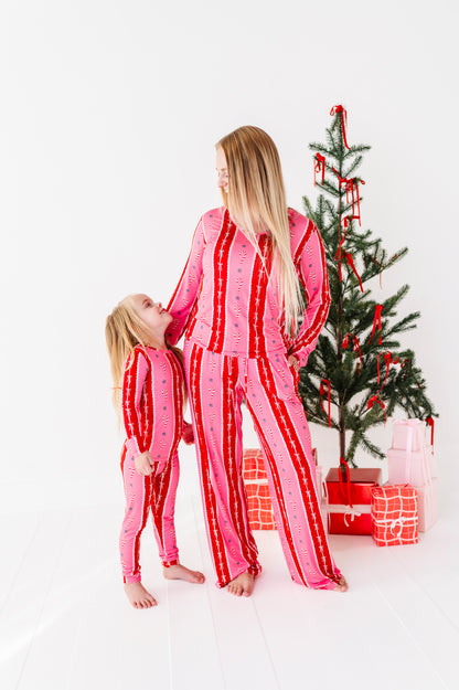 A woman and a child wearing matching pink and red striped bamboo pajamas pose in front of a decorated Christmas tree with wrapped gifts on a white studio floor.