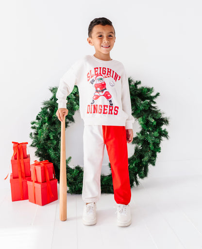 A young boy stands in a studio setting wearing a white sweatshirt and sweatpants set with a holiday theme. The sweatshirt features red collegiate-style lettering that reads 'SLEIGHIN' DINGERS' with a sleigh graphic, and the pants are split down the middle with one leg in white and the other in bright red.