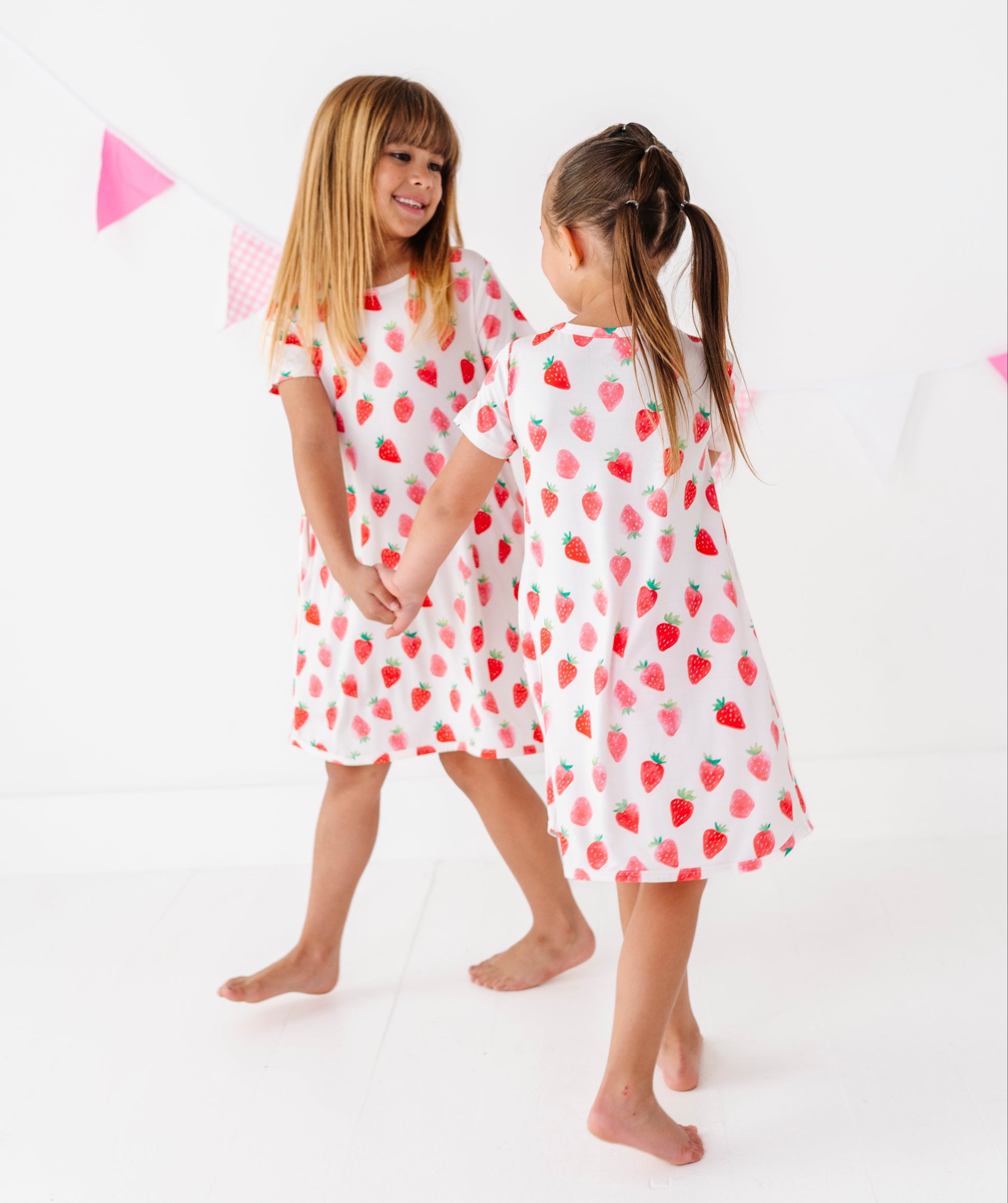 Two children wearing strawberry-patterned dresses standing on a white background with pink triangle flags.