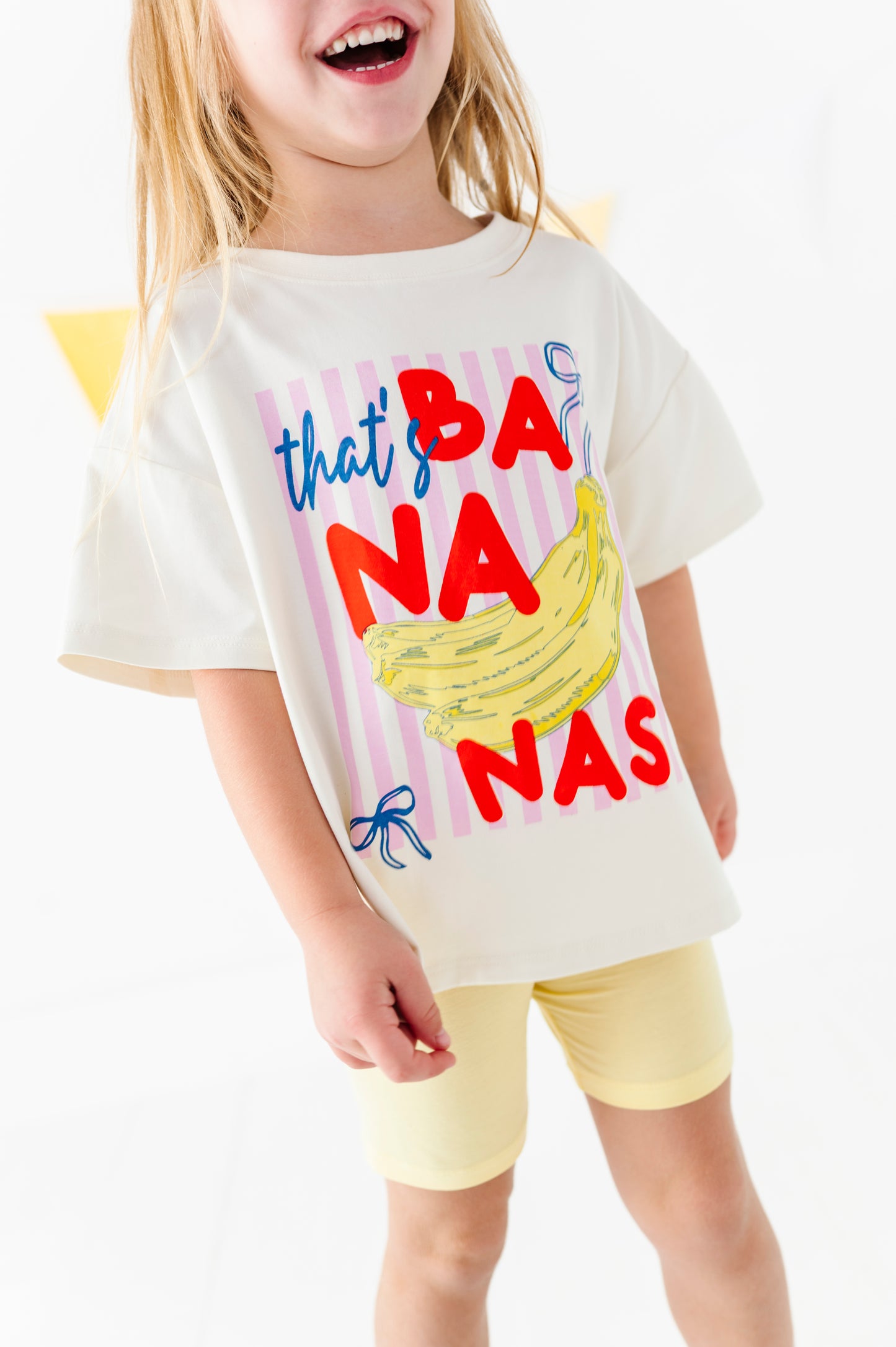 A young girl wearing a cream-colored graphic t-shirt with 'that's BANANA NAS' in red and blue text alongside a yellow banana illustration, paired with light yellow shorts.