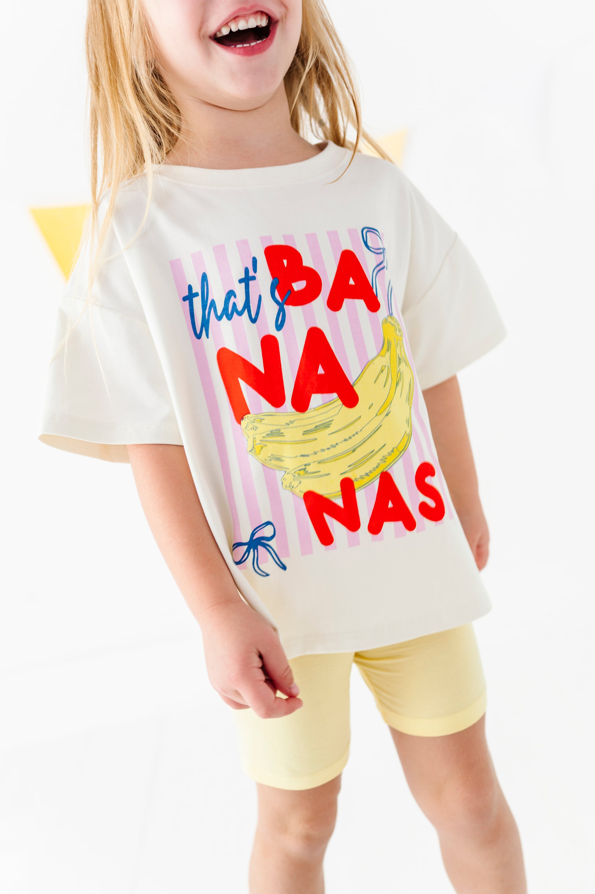 A young girl wearing a cream-colored graphic t-shirt with 'that's BANANA NAS' in red and blue text alongside a yellow banana illustration, paired with light yellow shorts.