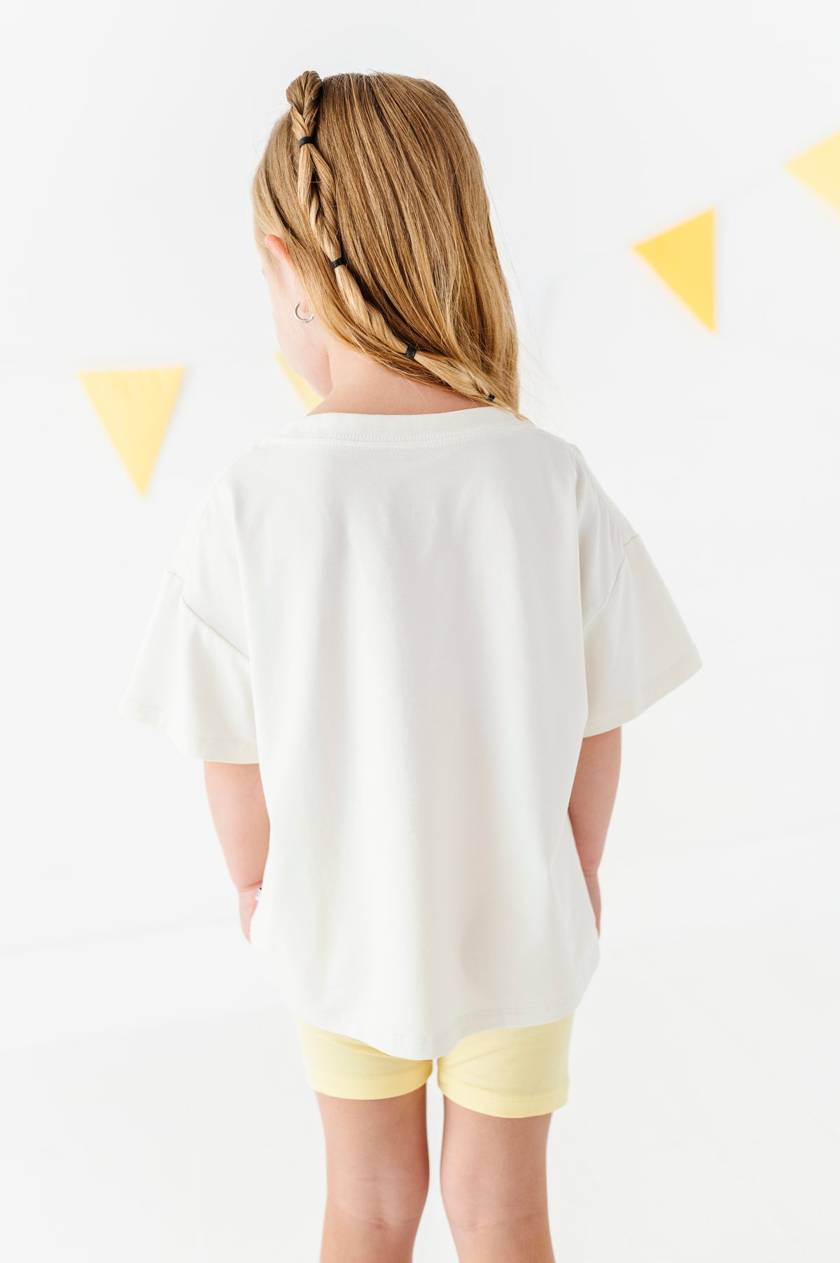 A child viewed from behind wearing an oversized off-white short-sleeve t-shirt and pale yellow shorts, standing against a white wall with yellow triangular bunting.