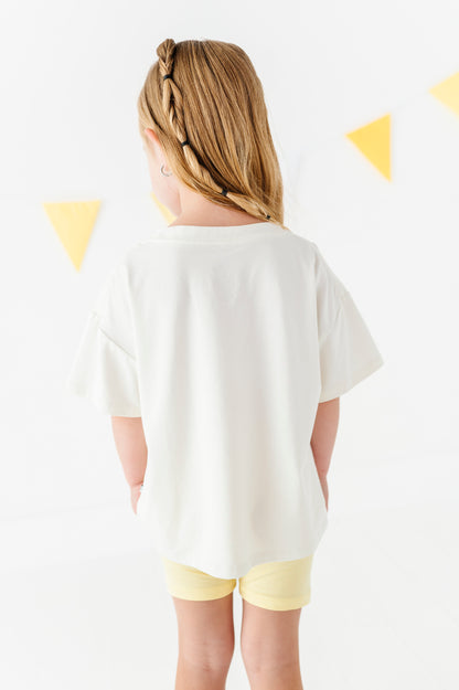 A child viewed from behind wearing an oversized off-white short-sleeve t-shirt and pale yellow shorts, standing against a white wall with yellow triangular bunting.