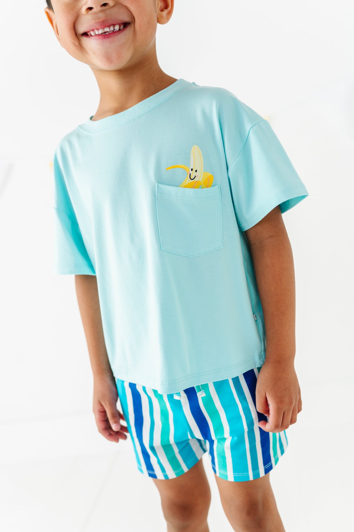 A smiling young boy wearing a light blue short-sleeve t-shirt with a yellow cartoon banana graphic in the chest pocket, paired with blue and white striped shorts, against a clean white background.