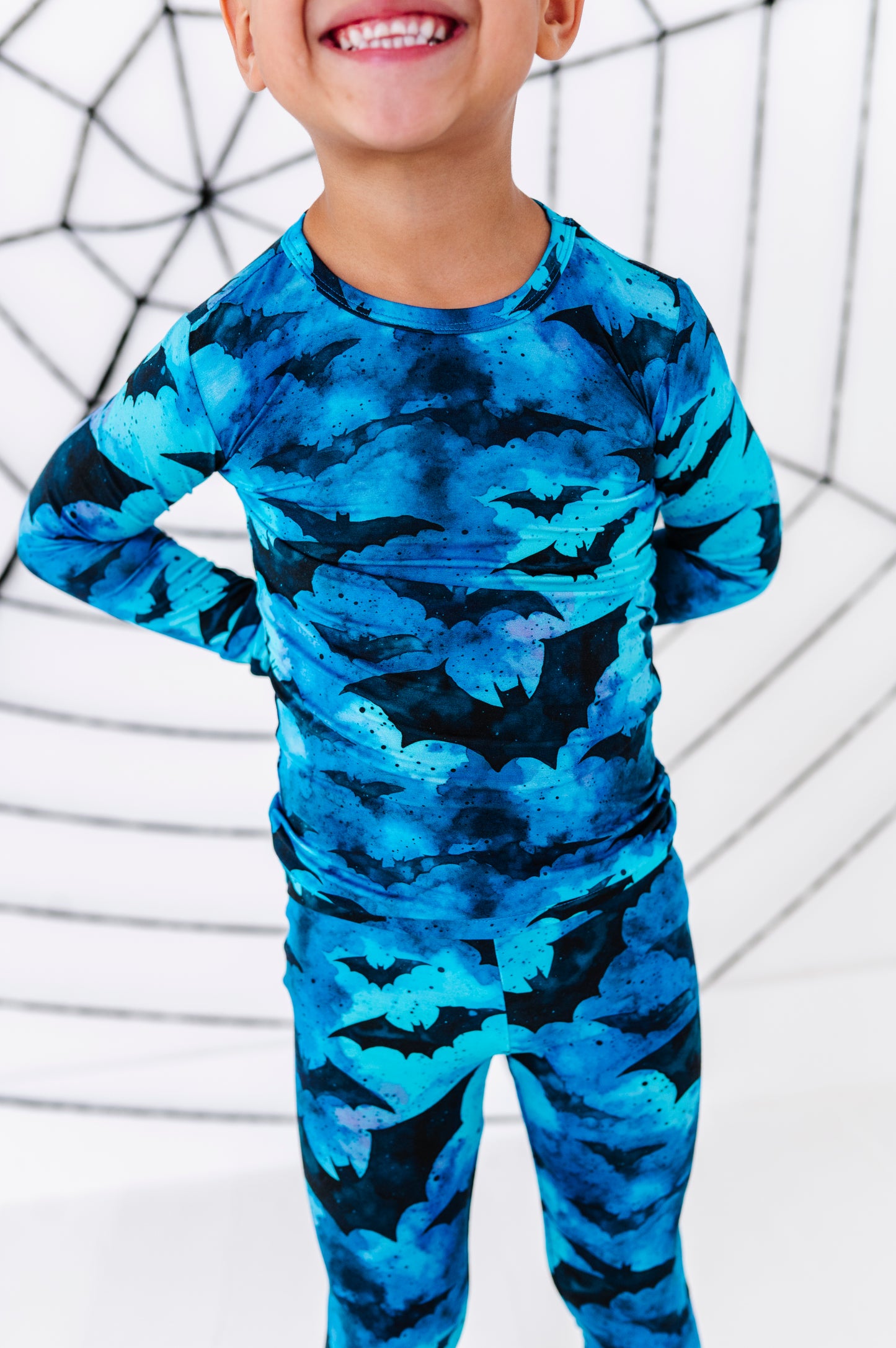 A child wearing a long-sleeve full-body onesie with a blue and black bat pattern, standing against a white background with black spiderweb graphics.