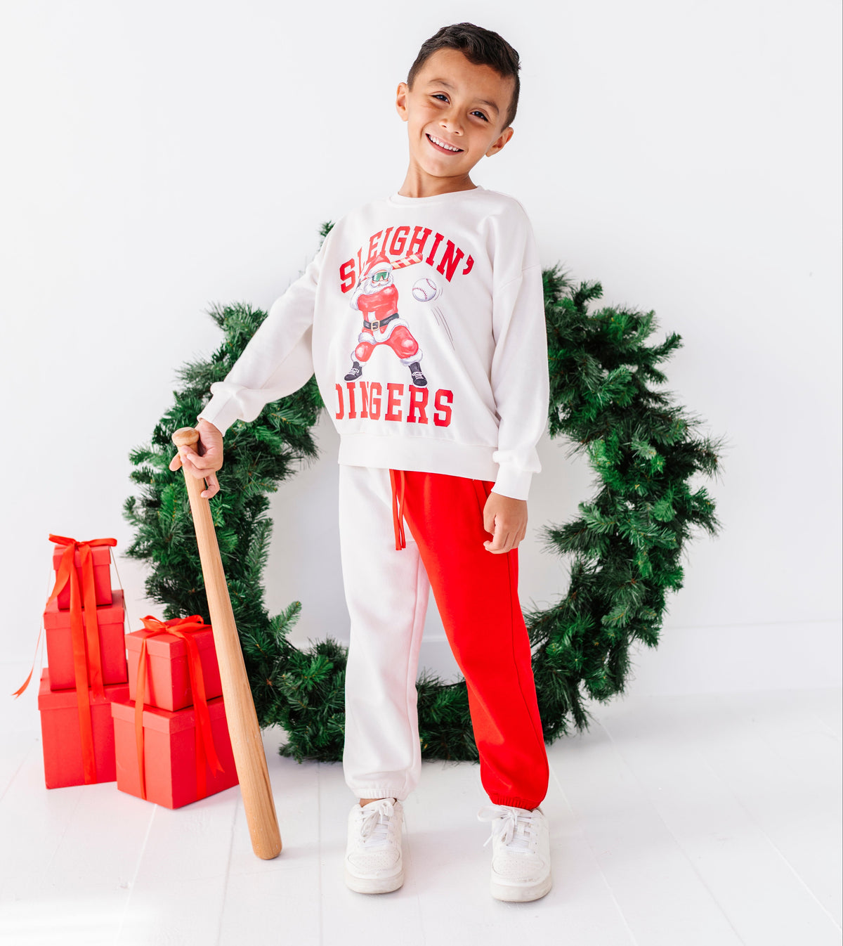 A young boy stands in a studio setting wearing a white long-sleeve sweatshirt with a red and blue 'Slighin' Tigers' graphic, paired with bright red sweatpants and white sneakers. He is holding a wooden baseball bat and posing in front of a green Christmas wreath with red gift boxes on the floor.