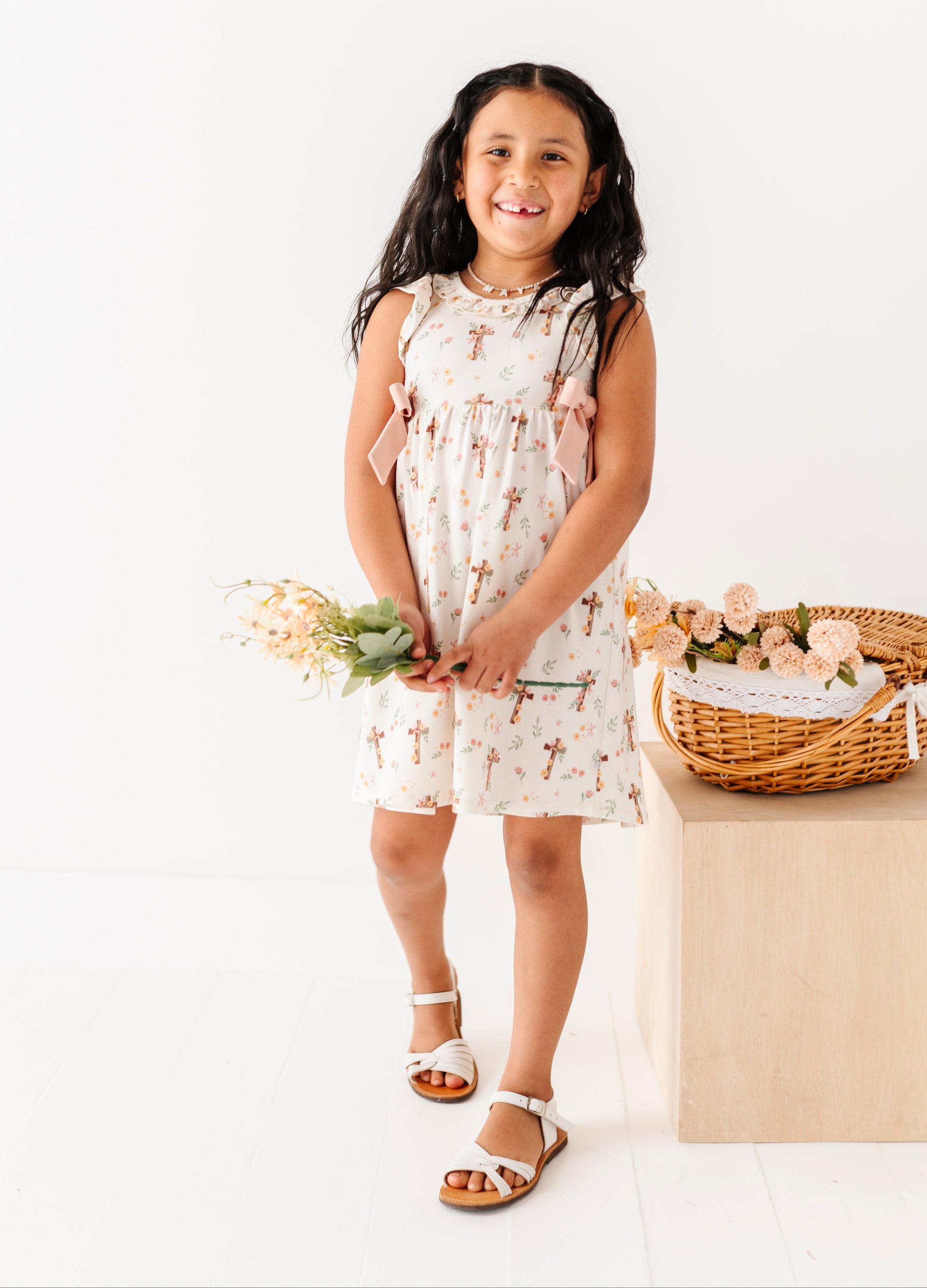 Young girl in a white dress with floral pattern holding flowers, standing next to a basket on a white background