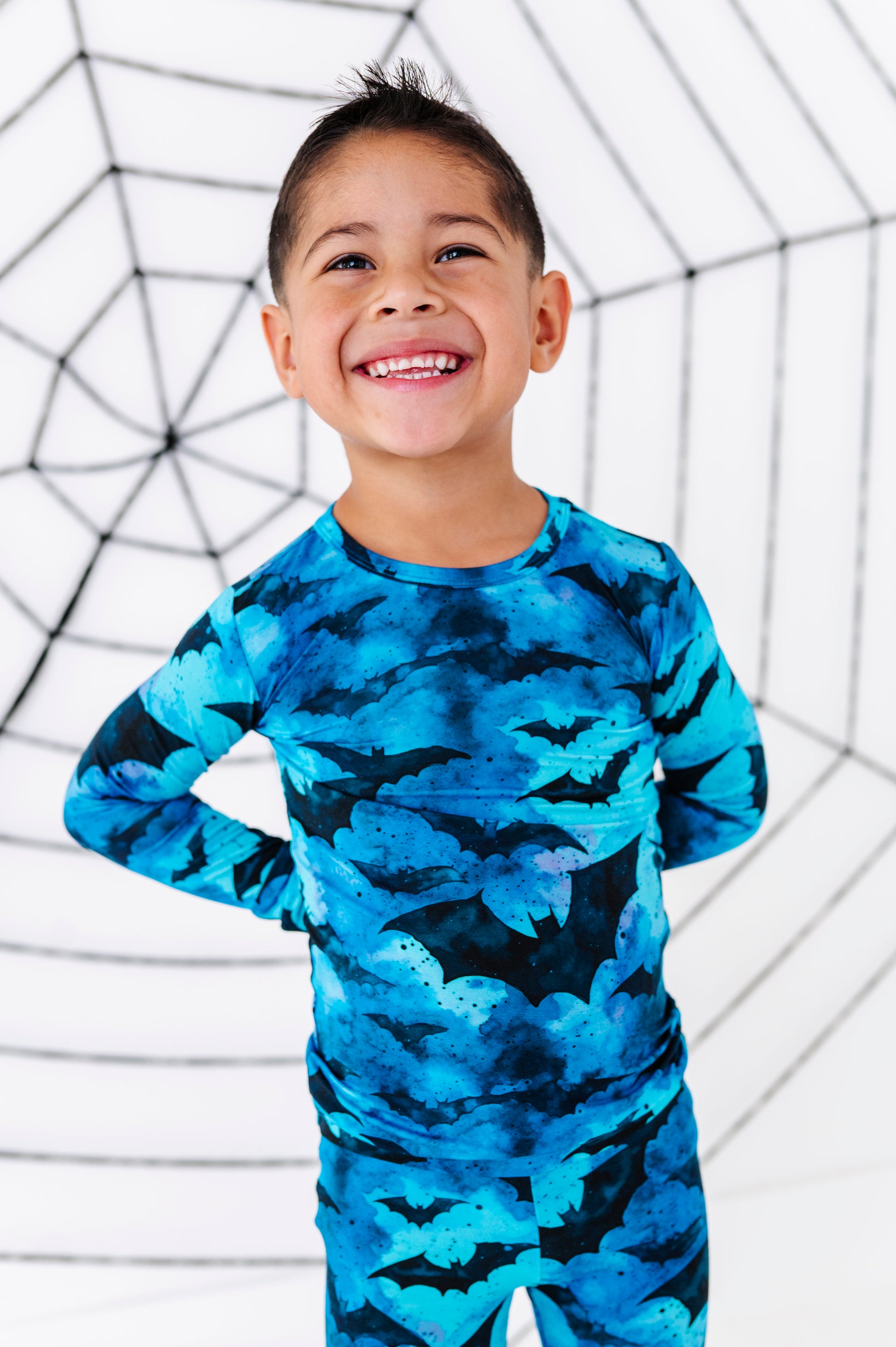 A young boy smiling and wearing a long-sleeve pajama set with a blue and black bat pattern, posed in front of a white background with a black spiderweb graphic.
