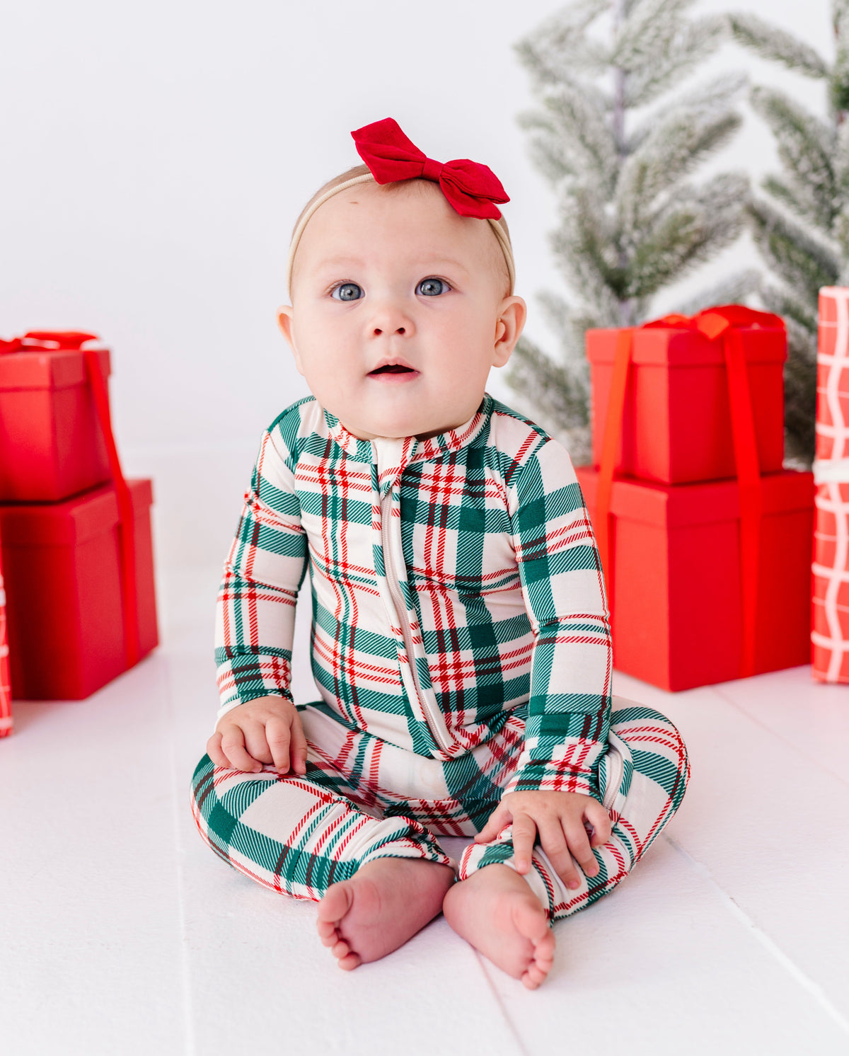 A baby sitting on a white floor wearing a green, red, and white plaid one-piece bamboo romper with a red bow in their hair, surrounded by red gift boxes and a snowy tree.