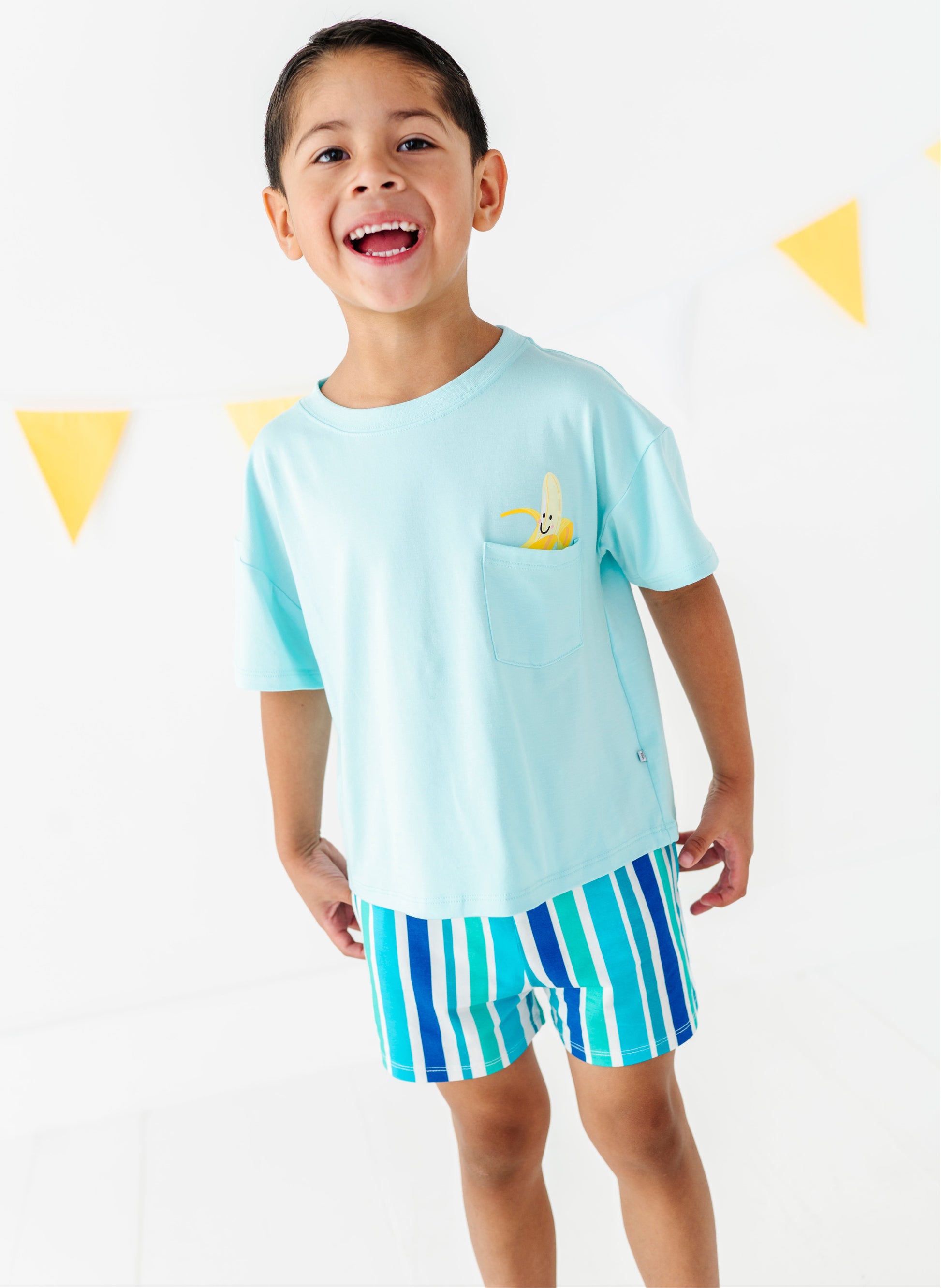 A young boy smiling and wearing a light blue short-sleeve t-shirt with a small yellow banana on the chest pocket, paired with blue and green striped shorts, set against a white wall with yellow triangular bunting.