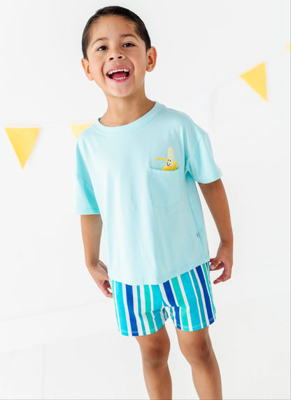A young boy smiling and wearing a light blue short-sleeve t-shirt with a small yellow banana on the chest pocket, paired with blue and green striped shorts, set against a white wall with yellow triangular bunting.