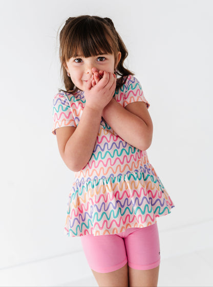 Young girl wearing a colorful wavy patterned bamboo/cotton peplum top and pink bamboo biker shorts on a white background